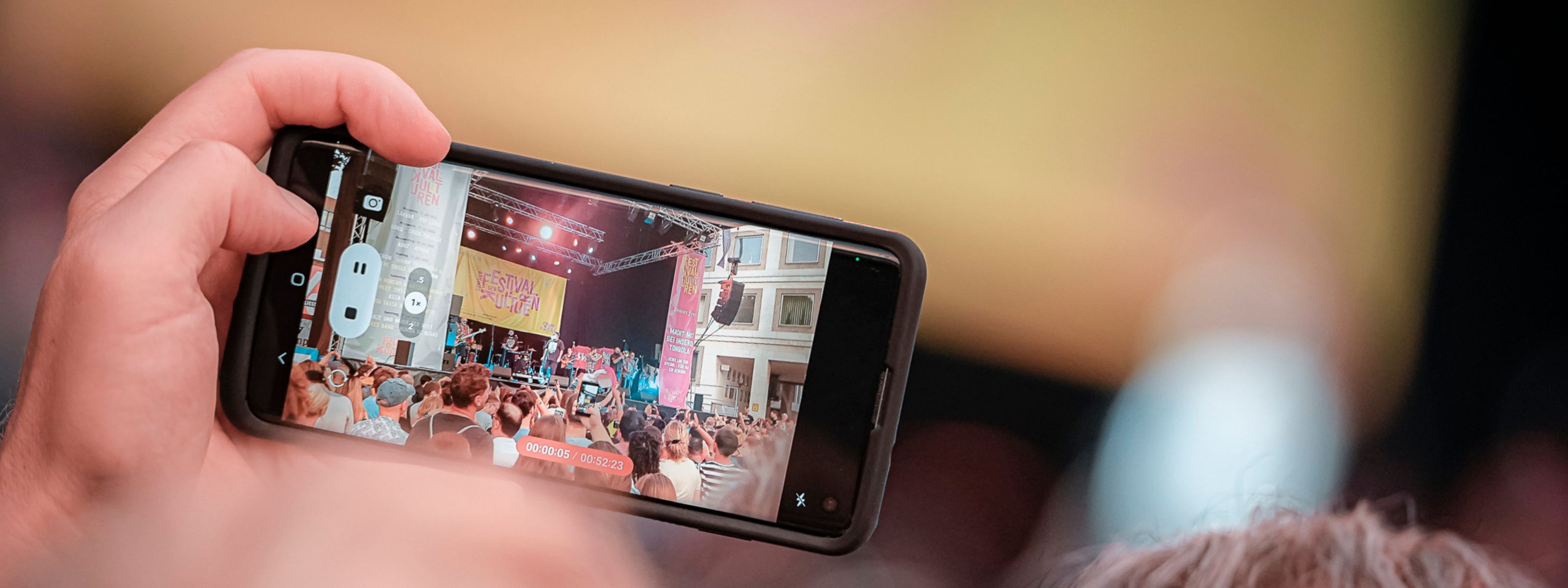 Das Bild zeigt eine Hand, die ein Handy hält und beim Sommerfestival der Kulturen filmt. Die Handykamera zeigt in Richtung Bühne. Auf dem Monitor des Handys sieht man eine Band, die auf der Bühne steht.