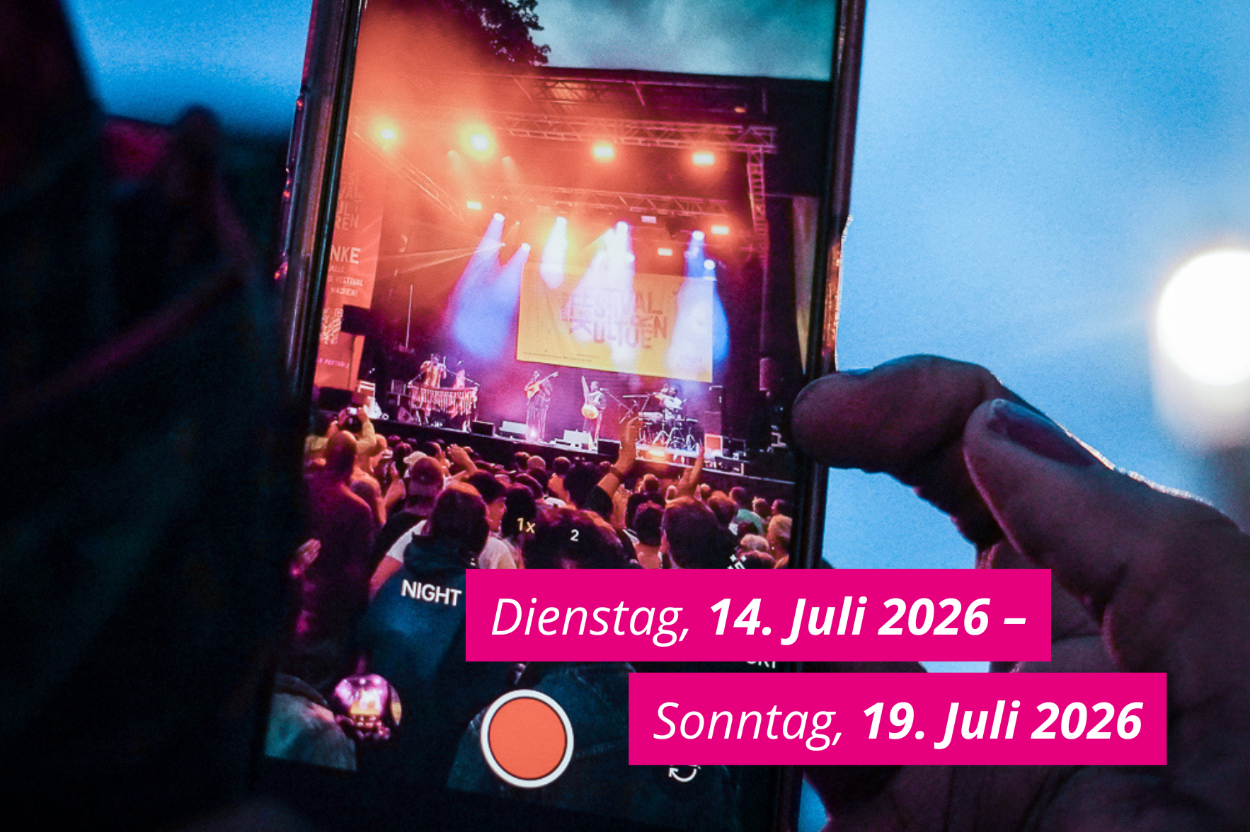 Das Bild zeigt ein Smartphone, das gerade in Richtung Bühne des Sommerfestivals der Kulturen filmt. Auf dem Bild ist ein pinkfarbener Balken, der das Datum des kommenden Festivals zeigt: Dienstag, 14. bis Sonntag, 19. Juli 2026